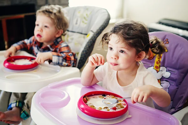 Portrait of cute adorable Caucasian children twins siblings sitting in high chair eating cereal early morning, everyday lifestyle candid moments, toned with Instagram filters — стоковое фото